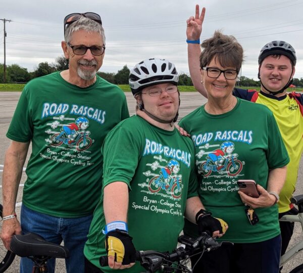 An older man and woman with their son wearing a cycling helmet in his late 20s wearing Road Rascals' cycling shirts and a young man standing behind them wearing a cycling helmet.